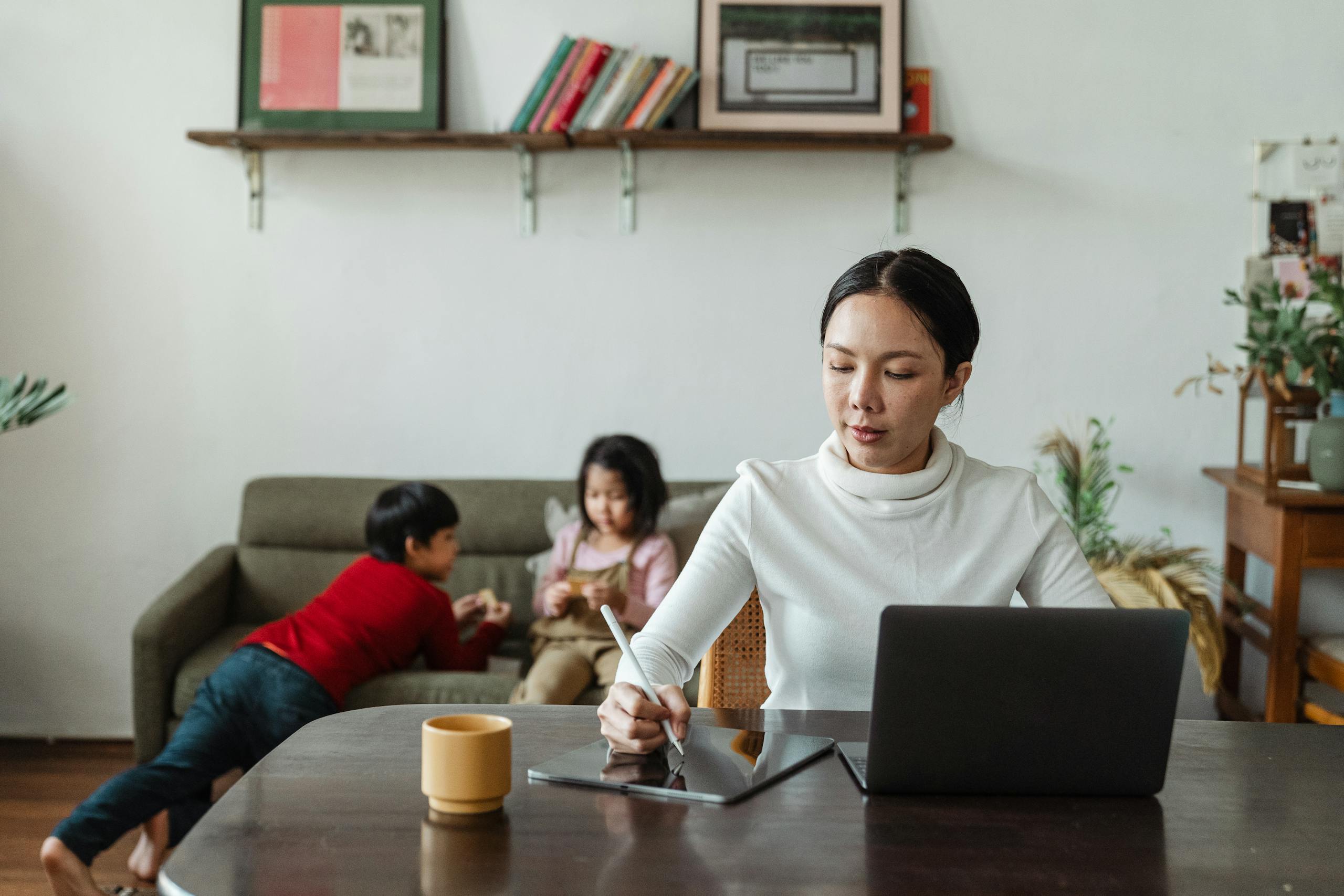 Mother multitasking with laptop and stylus alongside her children playing at home.